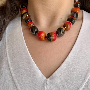 Puede incluir: Un collar grueso con grandes cuentas brillantes en tonos rojos, naranjas, marrones y negros. Las cuentas están ensartadas, creando una pieza audaz. El collar se lleva sobre una parte superior blanca.