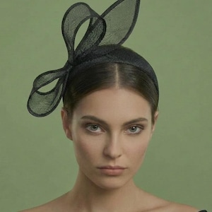 May include: A black headband with a decorative bow and a large, curved accent piece. The headpiece is made of a sheer, textured material. The woman in the image is wearing a black top against a green background.