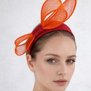 May include: A red headband with an orange bow detail. The bow is crafted from a sheer, textured material. The headband itself is a solid, deep red. This headpiece is suitable for special occasions.