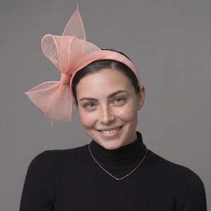 May include: A peach-colored headband featuring a large, structured bow made of sheer, stiffened fabric. The headband itself is covered in a matching peach-colored ribbed material, creating a cohesive look with the prominent, upright bow detail.