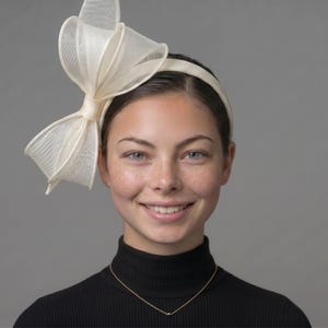 May include: An off-white fascinator headband featuring a large, structured bow made of stiffened, semi-sheer mesh fabric. The bow is positioned asymmetrically on the side of the thin, matching off-white headband, creating a sculptural effect.