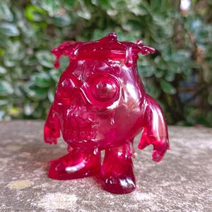 May include: A translucent red figurine of a creature with large eyes, a rounded body, and small arms and legs. The figurine has a textured surface and a dripping effect on its head and body. The background is blurred green foliage.