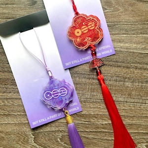 May include: Two decorative charms with tassels. One is a red cloud shape with a red tassel, the other is a purple flower shape with a purple tassel. Both have the text "BUT STILL A PART OF THIS WORLD".