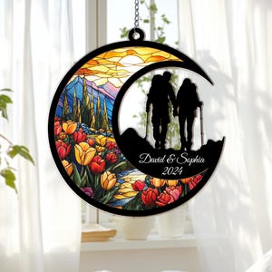 May include: A decorative crescent-shaped ornament featuring a stained-glass style landscape with mountains, tulips, and a couple hiking. The ornament includes the names "David & Sophia" and the year "2024".