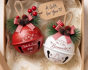 May include: Two round metal jingle bell ornaments in a gift box. One is red with 'Our First Home', 'Chole & Liam', and '2026' text. The other is white with 'First Christmas as Mr. & Mrs. Bennett', 'Chole & Liam', and '2026'. Both have pine, berries, and bows.