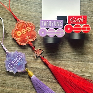 May include: A collection of decorative charms and name tags. The charms are in red and purple, with intricate designs and tassels. The name tags read "TAEHYUNG" and "SUGA" in white lettering. The items are arranged on a wooden surface.