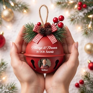 May include: A red metal jingle bell ornament featuring the text Always & Forever IN OUR hearts, a circular photo of a man, and the name William. The top is decorated with a red and white checkered bow, pine needles, pinecones, and red berries.