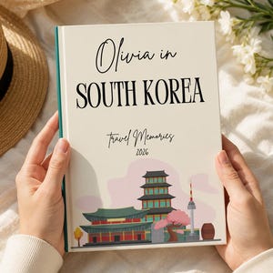 Pode incluir: Um livro de capa dura com uma capa cor de creme apresentando o texto Olivia in SOUTH KOREA Travel Memories 2026. A ilustração da capa retrata um edifício tradicional de vários níveis, uma torre alta e uma cerejeira em flor.