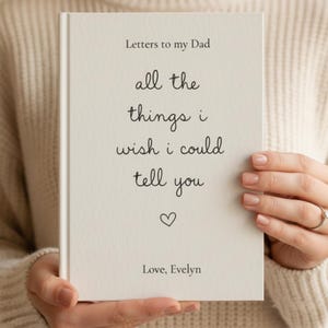 Può includere: Un libro color crema intitolato "Letters to my Dad" tenuto da una persona. La copertina presenta il testo "all the things i wish i could tell you" con un simbolo a cuore e "Love, Evelyn".