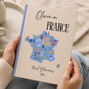 May include: A cream-colored travel journal titled "Olivia in France" with a blue map of France. The cover features illustrations of landmarks and travel icons. The words "Travel Memories 2026" are printed at the bottom.