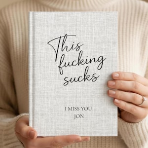 May include: A light gray journal with the text "This fucking sucks" in black script. Below, the text reads "I miss you Jon". The journal is held by a person wearing a cream-colored sweater.