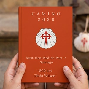 以下が含まれることがあります： 「CAMINO 2026」の文字とホタテのデザインが施された赤いジャーナル。 「Saint-Jean-Pied-de-Port -> Santiago」と「~800 km Olivia Wilson」の文字も表示されています。 ジャーナルは誰かの手に持たれています。