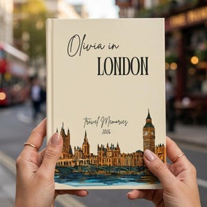 May include: A cream-colored travel journal titled "Olivia in London" with a London skyline illustration. The cover also includes the text "Travel Memories 2026". The journal is held in front of a blurred street scene.