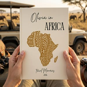 Op de afbeelding: Een crèmekleurig reisdagboek getiteld "Olivia in AFRICA" met een goudkleurige kaart van Afrika. De woorden "Travel Memories" en het jaar "2026" staan op de omslag. Het dagboek wordt voor een safarivoertuig gehouden.