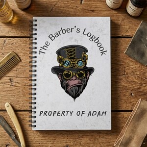 May include: A spiral-bound notebook with the title "The Barber's Logbook" and the text "Property of Adam." The cover features a steampunk-style illustration of a monkey wearing a top hat and goggles. The notebook is on a wooden surface.