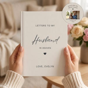 May include: A cream-coloured book titled "Letters to My Husband in Heaven" with a black heart and the name "Evelyn" at the bottom. A small circular photo on the back cover is visible. The book is held by a person.