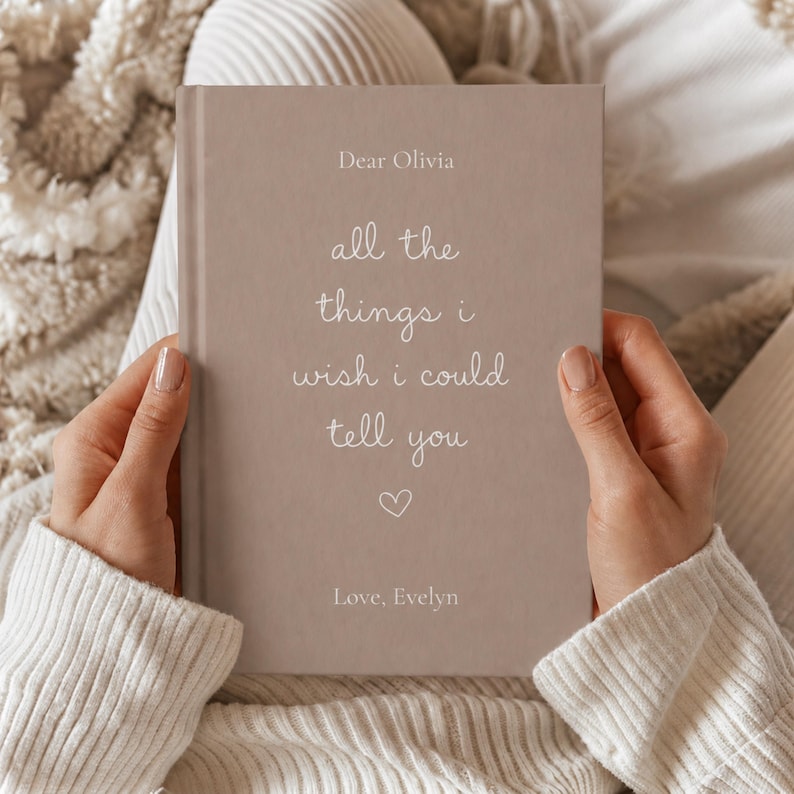 Może przedstawiać: Notatnik w twardej oprawie w kolorze taupe z białym napisem Dear Olivia, all the things i wish i could tell you, małą ikoną serca oraz Love, Evelyn. Książka jest trzymana pionowo, prezentując przednią okładkę o matowym wykończeniu.