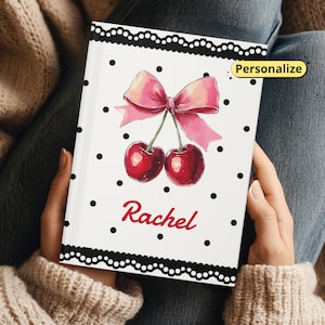 Pode incluir: Um caderno branco com um laço rosa e uma ilustração de duas cerejas vermelhas. O caderno tem bolinhas pretas e uma borda de renda preta. O nome "Rachel" está escrito em vermelho. A palavra "Personalizar" está em uma caixa amarela.