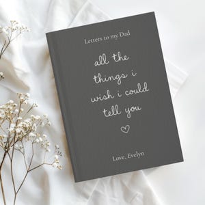 Może przedstawiać: Ciemnoszary dziennik w twardej oprawie z białym napisem Letters to my Dad, all the things i wish i could tell you, małą ikoną serca oraz imieniem Love, Evelyn nadrukowanym na przedniej okładce wyraźną czcionką oraz pismem odręcznym.