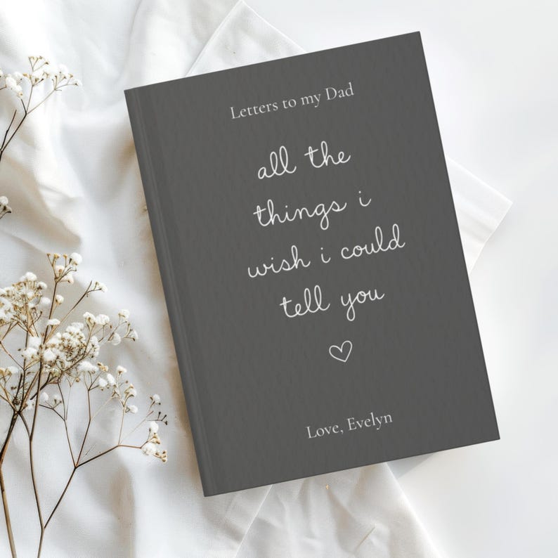 Może przedstawiać: Ciemnoszary dziennik w twardej oprawie z białym napisem Letters to my Dad, all the things i wish i could tell you, małą ikoną serca oraz imieniem Love, Evelyn nadrukowanym na przedniej okładce wyraźną czcionką oraz pismem odręcznym.