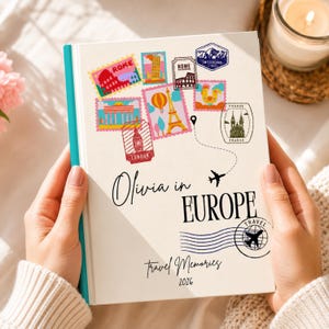 Puede incluir: Un diario de viaje color crema con lomo turquesa, sostenido por manos. La portada presenta diseños de sellos postales con temática de viajes con monumentos de Roma, Londres y otras ciudades europeas. El texto dice "Olivia en Europa" y "Recuerdos de viaje 2026."