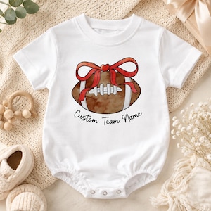 May include: A white short-sleeved baby bodysuit featuring a brown football graphic with a red bow and a small red heart on top. The text Custom Team Name is printed in black cursive script below the football illustration on the front of the garment.