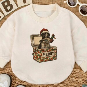 Könnte beinhalten: Cremefarbener Baby-Body mit langen Ärmeln. Auf der Vorderseite befindet sich eine Grafik eines Hundes mit Weihnachtsmütze, der eine Ente hält und in einem Camouflage-Kühler mit dem Wort "MERRY" sitzt. Weiße Babyschuhe und Sonnenbrille.