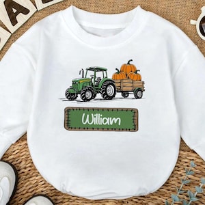 May include: White baby long-sleeve shirt with a green tractor pulling a wagon of pumpkins. The name "William" is printed on a green rectangle. The word "BABY" is spelled out in wooden blocks.