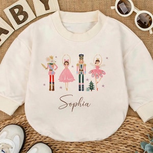 May include: Cream-colored baby bodysuit with a Nutcracker-themed graphic. The design features four characters in colorful outfits, a Christmas tree, and the name "Sophia" in script. White baby shoes and sunglasses are also in the image.