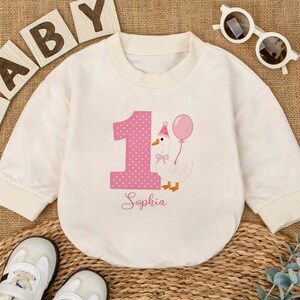 May include: A cream-colored long-sleeve sweatshirt featuring a large pink polka-dot number one, a white goose wearing a pink party hat and holding a pink balloon, and the name Sophia printed in cursive script below the graphic.