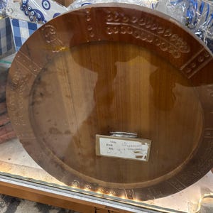 May include: A round, wooden serving tray with a dark brown finish. The tray has carved decorative patterns around the rim. A small label is attached to the tray.