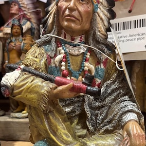 May include: A detailed figurine of a Native American chief, seated and holding a peace pipe. The figure wears a feathered headdress, beaded necklaces, and traditional clothing in shades of yellow, blue, and red. The figurine has a weathered appearance.