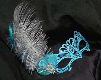 May include: A blue masquerade mask with a delicate, ornate design. The mask is adorned with rhinestones and a single gray feather.