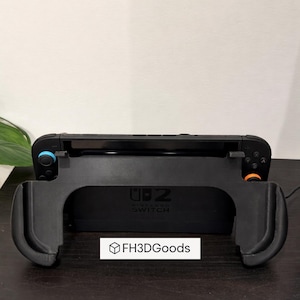 Può includere: Console Nintendo Switch nera con un'impugnatura nera stampata in 3D. L'impugnatura ha maniglie curve e il testo "UB 2 Nintendo Switch". È visibile anche il logo "FH3DGoods".