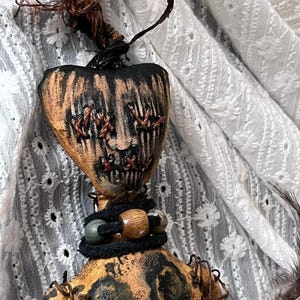 May include: A handmade Halloween-themed doll with a heart-shaped head and a stitched face. The doll features a black and orange color scheme, with a textured, distressed appearance. It is adorned with beads and a dark, twisted stem.