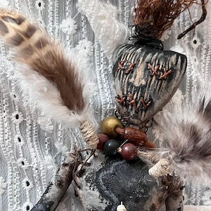May include: A handmade doll with a heart-shaped head and stitched facial features. The doll has a patterned body, adorned with feathers, beads, and natural elements. The color palette includes black, brown, and cream.