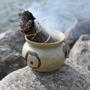 Può includere: Un fascio di salvia che brucia in una piccola ciotola di ceramica verde chiaro con decorazioni marroni. La salvia è legata con uno spago bianco e il fumo si alza dall'estremità in fiamme. La ciotola è su una grande roccia grigia.