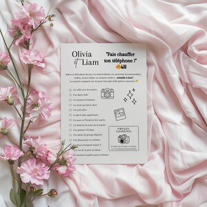 Pu&ograve; includere: Una carta bianca con i nomi "Olivia et Liam" e la frase "Fais chauffer ton t&eacute;l&eacute;phone!" in testo nero. La carta include un elenco di idee per foto di matrimonio, con illustrazioni di una fotocamera, cuori e stelle. Fiori e tessuto rosa circondano la carta.