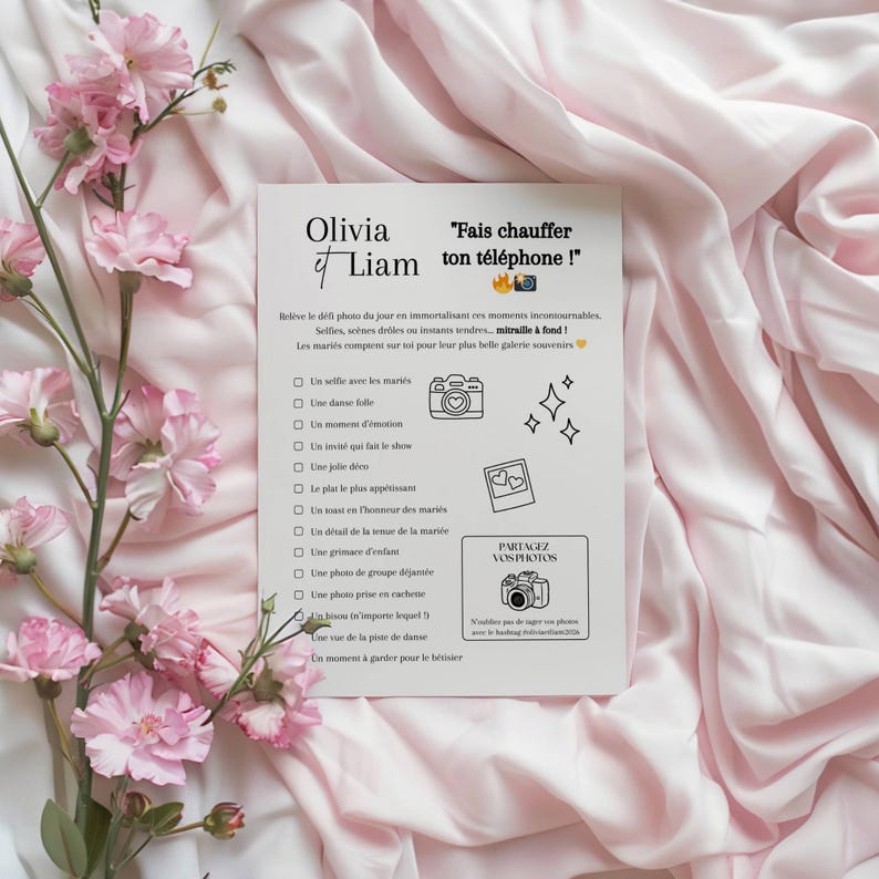 Pu&ograve; includere: Una carta bianca con i nomi "Olivia et Liam" e la frase "Fais chauffer ton t&eacute;l&eacute;phone!" in testo nero. La carta include un elenco di idee per foto di matrimonio, con illustrazioni di una fotocamera, cuori e stelle. Fiori e tessuto rosa circondano la carta.