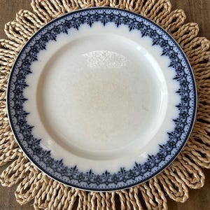 May include: A white ceramic plate with a blue floral border sits on a woven placemat. The plate has a simple, elegant design, with a delicate blue pattern around the rim. The placemat is made of natural fibers and adds a rustic touch.