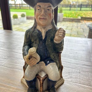 May include: A ceramic figure of a man in a tricorn hat, dark blue coat, and white breeches, seated on a chair. The figure holds a money bag and clenches a fist. The figurine is painted in muted tones.