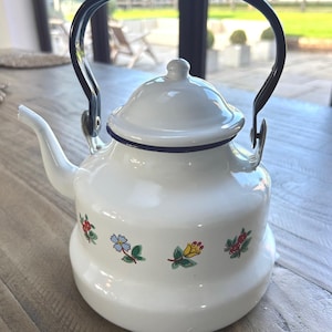 Puede incluir: Tetera de esmalte blanco con asa y boquilla negras. La tetera tiene tapa y está decorada con diseños florales coloridos. El asa es arqueada y la boquilla es curva. Un artículo de cocina clásico.