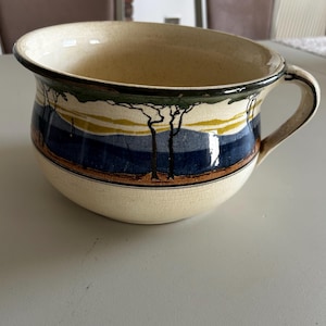 May include: Cream-colored ceramic bowl with a handle and a landscape design. The bowl features a dark blue band with a yellow and green horizon line, and black tree silhouettes. The rim is dark brown.