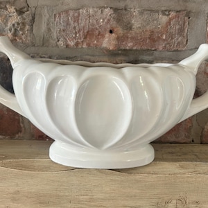 Może przedstawiać: Biała ceramiczna misa z falistym, przypominającym płatki wzorem na zewnątrz oraz dwoma zakrzywionymi uchwytami po przeciwnych stronach. Misa spoczywa na okrągłej podstawie i ma gładkie, błyszczące wykończenie na całej swojej powierzchni.