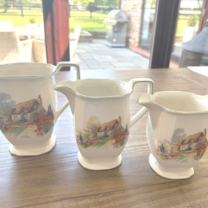 May include: Three white ceramic pitchers with a cottage scene design. Each pitcher has a handle and a spout. The cottage scene features a thatched roof house, trees, and flowers. The pitchers are arranged on a wooden surface.