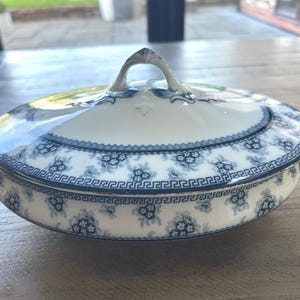Op de afbeelding: Ovale serveerschaal met deksel, met een wit en blauw bloemmotief. De schaal heeft een handvat op het deksel en twee handvatten aan de zijkanten. De rand heeft een Grieks sleutelpatroon. Een klassiek stuk om eten te serveren.