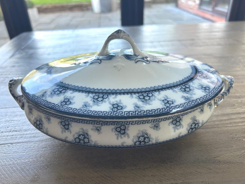 Op de afbeelding: Ovale serveerschaal met deksel, met een wit en blauw bloemmotief. De schaal heeft een handvat op het deksel en twee handvatten aan de zijkanten. De rand heeft een Grieks sleutelpatroon. Een klassiek stuk om eten te serveren.