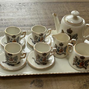 May include: A vintage tea set on a white tray with gold trim. The set includes a teapot, creamer, sugar bowl, and six teacups with saucers. Each piece features a crest design and gold accents.