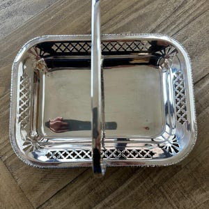 Puede incluir: Una bandeja rectangular plateada con asa. La bandeja tiene un diseño calado decorativo alrededor de los bordes y una superficie pulida y reflectante. El asa está centrada y en posición vertical.