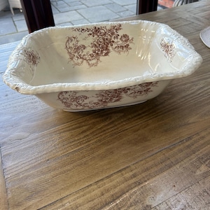 Peut inclure: Bassin rectangulaire en céramique crème avec un motif floral décoratif dans les tons bruns. Le bassin a une bordure texturée en forme de corde et est posé sur une surface en bois. Le motif floral se répète à l'intérieur du bassin.